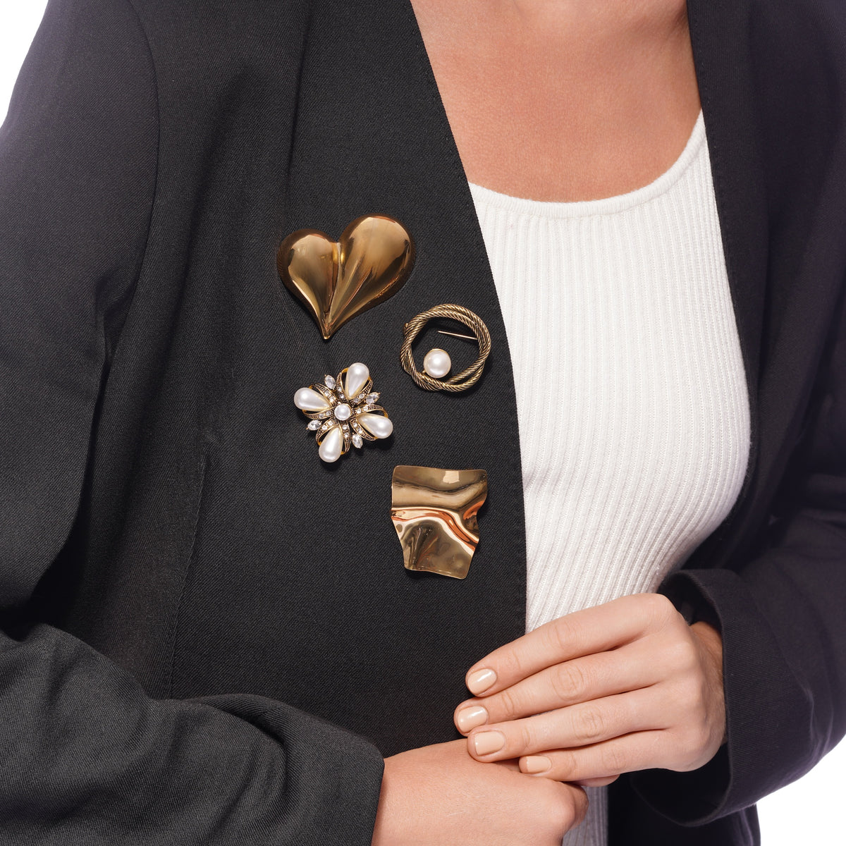 A close-up of a person wearing a black blazer over a white ribbed top, styled with four distinct vintage-style brooch pins on the left side of the blazer.