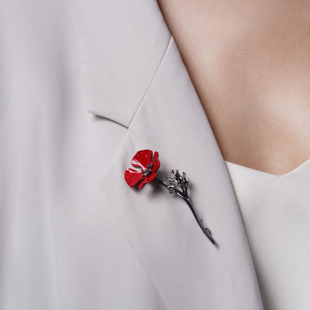 Red enamel poppy brooch with a dark metallic stem worn diagonally on a light blazer lapel.
