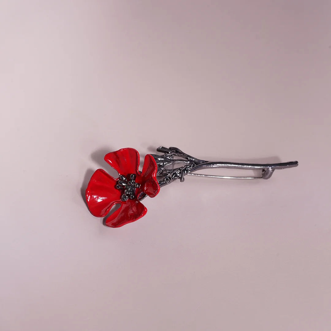 Red enamel poppy brooch laid horizontally, angled view showing the petals, metal stem and pin.